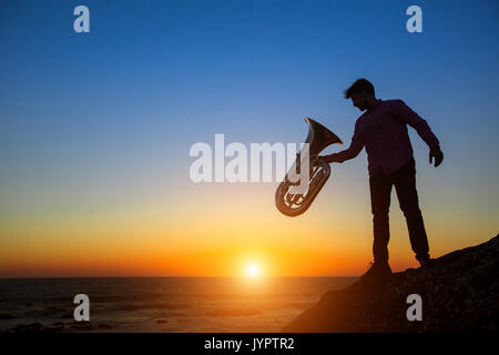 Silhouette des Musikers mit Tuba Instrument auf felsigen Küste bei Sonnenuntergang. Stockfoto