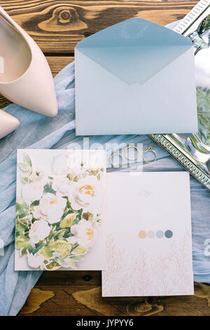 Zubehör, Ehevertrag, Zärtlichkeit Konzept. Herrliche flatlay von Pastelltönen. Es ist hellblau Kuvert Brief, zwei Postkarten mit floralem Design. Drei goldene Ringe und rosa Schuhe der Braut. Stockfoto