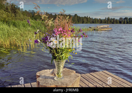 Schneiden Sie die wilden Blumen in einer Glasvase am Pier, einen Wald und See. Stockfoto