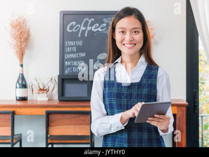 Asiatische Frau barista tragen blaue Schürze halten Tablet Computer/Menü mit lächelnden Gesicht im Cafe warten um von Kunden, Kaffee Geschäft Inhaber con Stockfoto