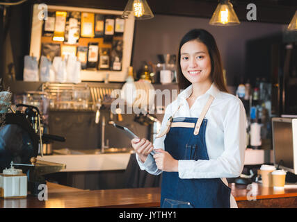 Asiatische Frau barista Verschleiß jean Schürze halten Tablet Computer/Menü counter Bar mit einem Lächeln im Gesicht, cafe Service Konzept. Stockfoto