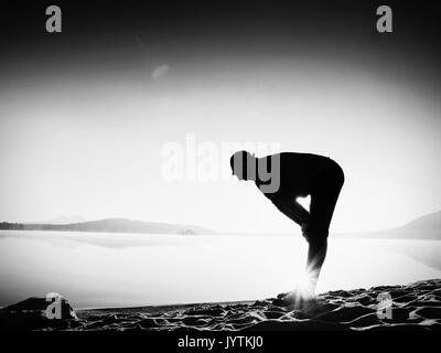 Tiefes Atmen runner Silhouette mit den Händen auf die Knie gebeugt. Seitenansicht zu Sport aktiven Mann stretching Muskeln und Bewegung am Meer Strand. Sun spiegeln Stockfoto