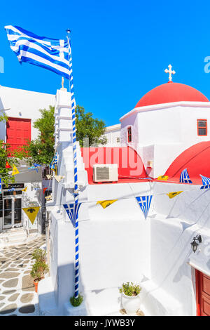 Insel Mykonos, Griechenland - 17. MAI 2016: Kirche Gebäude mit rotem Dach und die griechische Flagge auf der weiß getünchten Straße in der schönen Stadt Mykonos, Kykladen, Stockfoto