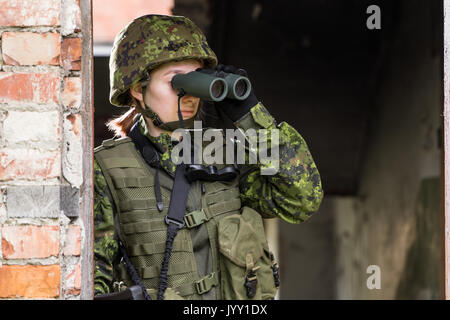 Portrait von bewaffneten Frau mit Camouflage. Junge weibliche Soldaten mit dem Fernglas beobachten. Kind Soldat mit Gewehr im Krieg, Haus Ruinen Hintergrund. Militar Stockfoto