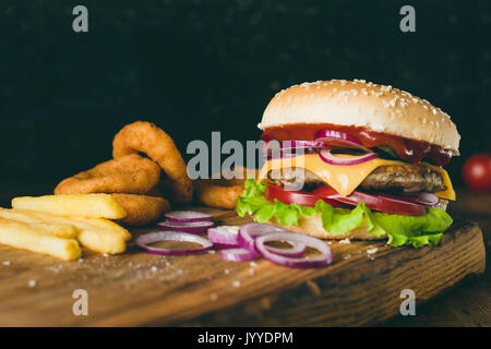 Cheeseburger, Pommes frites und Zwiebelringe auf Holz Schneidebrett über Holz- Hintergrund. Detailansicht, selektive konzentrieren. Fast food Konzept Stockfoto