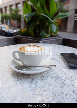 Herzförmige Kunst auf einen Cappuccino mit einem Telefon auf einem Marmortisch Stockfoto