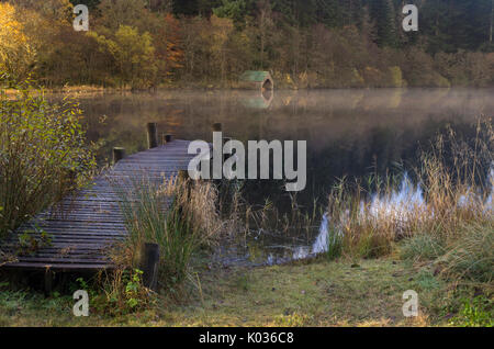 Holzsteg und Bootshaus auf einem nebligen Morgen mit Reflexion, Loch Ard, Schottland Stockfoto