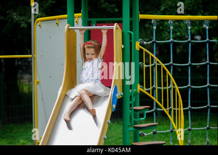 Ein süßes kleines Barfuß girly ist das Spielen auf dem Spielplatz. Baby sitzt auf die Kinder schieben. Auf den Kopf des Mädchens ist ein roter Kranz. Stockfoto