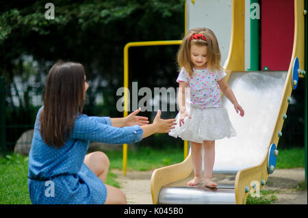 Mutter und ihre kleine Tochter Spielen auf dem Spielplatz zu. Barfuß Baby steht auf der Kante der Folie. Mom streckt die Hand nach dem Mädchen. Stockfoto