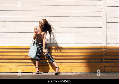 Outdoor portrait einer jungen schönen modischen Happy lady Sonnenbrille tragen, in einem Holzrahmen, gegen eine Holz-, weiße Wand. Das Model trägt stilvolle Kleidung, eine beige Bluse, schwarzen Hosen in einen weißen Streifen, eine Tüte mit Hellblau. Das Mädchen schaut weg und Getränke rosa Mors und Rohre. Damenmode. Städtische Lebensweise Stockfoto