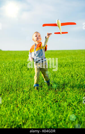 Fröhlicher Junge spielt mit Spielzeugflugzeug gegen blauen Himmel und grüne Feld Hintergrund. Stockfoto