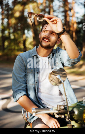 Foto von hübscher junger bärtiger Mann sitzen auf Roller im Freien holding Sonnenbrille. Neben der Suche. Stockfoto