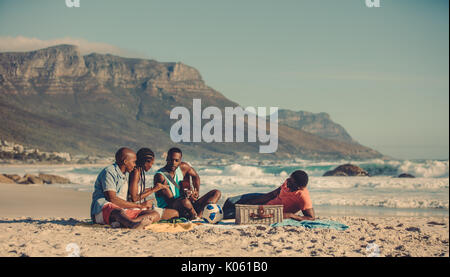 Afrikanischer Mann Gitarre spielen für seine Freunde entlang der Küste. Gruppe von Menschen Entspannung am Sandstrand. Stockfoto