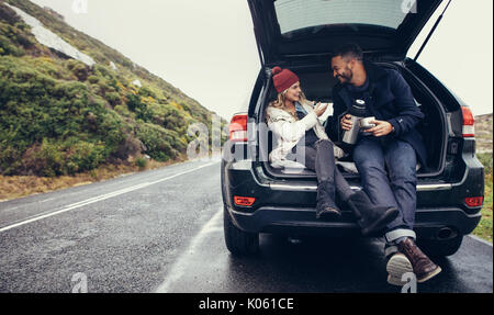 Glückliches junges Paar in einer Kaffeepause während der Fahrt in die Landschaft. Mann und Frau im Kofferraum sitzen und Kaffee. Stockfoto