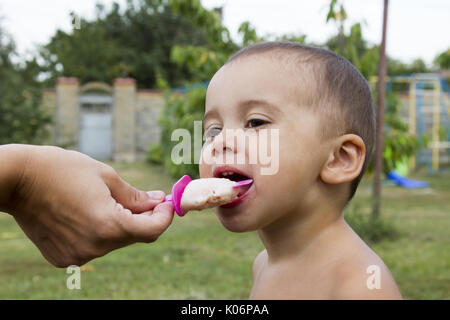 Süßes Kleinkind Baby junge achtzehn Monate alt, leckeres Eis über Hintergrund mit grünen Bokeh im Freien zu essen. Stockfoto