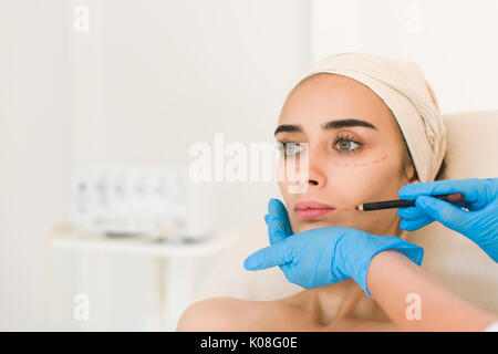 Attraktives Mädchen mit dunklen Augenbrauen, die Hände des Arztes trägt blaue Perforationslinien Handschuhe Zeichnung im Gesicht. Stockfoto