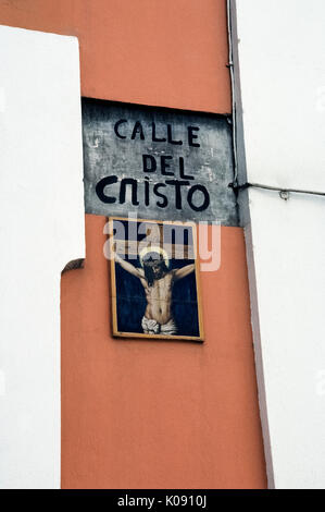 Eine religiöse Kunstwerke von keramischen Fliesen zeigt Jesus Christus am Kreuz und markiert die Calle del Cristo (Straße, in der sich das Kruzifix), die zu der Kathedrale von San Juan Bautista im historischen alten San Juan in Puerto Rico (PR), ein gemeindefreies Gebiet der Vereinigten Staaten in der Karibik Meer führt. Ein vintage Schmiedeeisen Straße Licht der Straße Name aufleuchtet und illustrierte Zeichen in der Nacht. Stockfoto