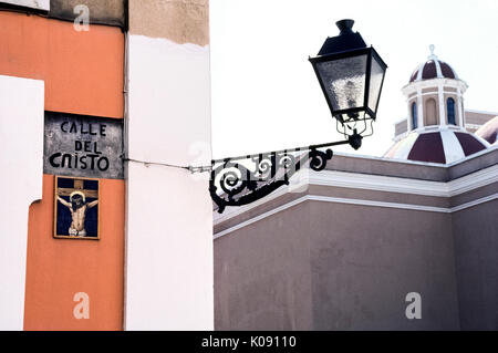 Eine religiöse Kunstwerke von keramischen Fliesen zeigt Jesus Christus am Kreuz und markiert die Calle del Cristo (Straße, in der sich das Kruzifix), führt zu der Kathedrale von San Juan Bautista (rechts) in der historischen Altstadt San Juan in Puerto Rico (PR), ein gemeindefreies Gebiet der Vereinigten Staaten in der Karibik. Ein vintage Schmiedeeisen Straße Licht der Straße Name aufleuchtet und illustrierte Zeichen in der Nacht. Stockfoto