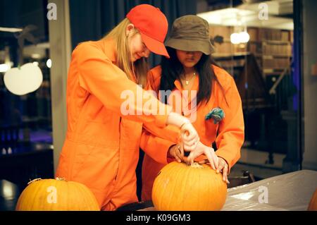 Zwei Studenten zusammen bei Carving die Stammzellen aus einem Kürbis arbeiten, sie sind in orange Overalls als Charaktere aus der Geschichte 'Löcher', Halloween an der Johns Hopkins University's George Peabody Library, 2015 bekleidet. Mit freundlicher Genehmigung von Eric Chen. Stockfoto