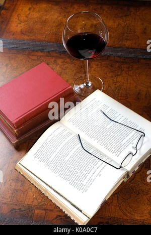 Ein offenes Buch auf einem Leder Couchtisch sitzt neben einem Stapel Bücher und einem Glas Rotwein. Lesebrille auf dem geöffneten Seiten. Stockfoto