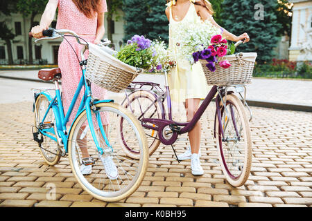 Zugeschnittenes Bild von zwei jungen Frauen in Kleidern mit retro Fahrrad auf einer Straße der Stadt Stockfoto