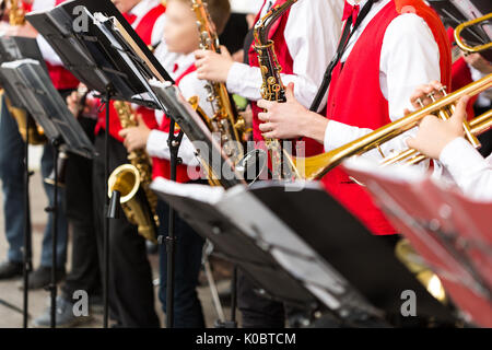 Musical Instrument, Brass Band und Orchester Konzept: Detailansicht Ensemble Musiker spielen auf Trompete und Saxophon in rot Konzert Kostüme, Notenständer, männlichen Händen mit Ausrüstung, selektiven Fokus. Stockfoto