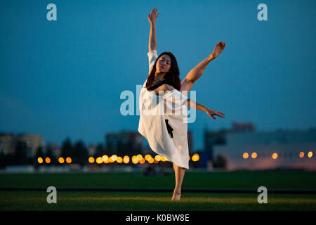 Ballerina in weißem Kleid steht und führt Schlucken auf Rasen und Gebäude Hintergrund in Abend darstellen. Nacht leuchten in der Ferne sichtbar. Stockfoto