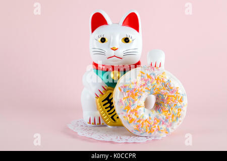 Ein Maneki Neko präsentiert eine mehrfarbige Donuts auf einem deckchen Papier und ein Pop-farbigen Hintergrund. Minimale schrulligen Farbe still life Fotografie Stockfoto