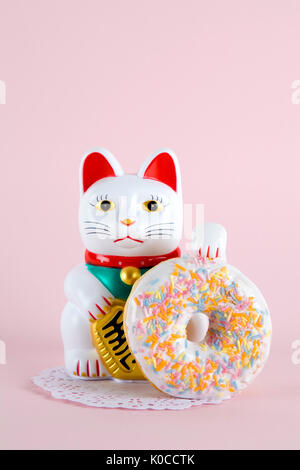 Ein Maneki Neko präsentiert eine mehrfarbige Donuts auf einem deckchen Papier und ein Pop-farbigen Hintergrund. Minimale schrulligen Farbe still life Fotografie Stockfoto