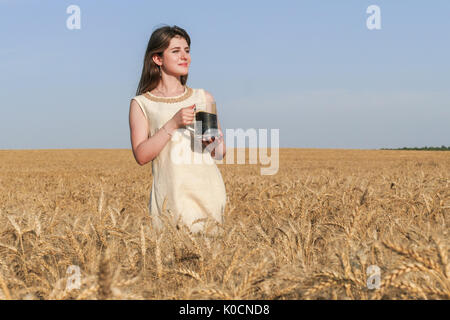 Junge attraktive Frau in wunderschönen natürlichen Kleid wandern mit mit einem Glas Bier in der goldene Weizen Feld während der Sonnenschein. Stockfoto