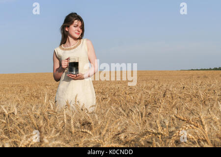 Junge attraktive Frau in wunderschönen natürlichen Kleid wandern mit mit einem Glas Bier in der goldene Weizen Feld während der Sonnenschein. Stockfoto