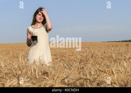 Junge attraktive Frau in wunderschönen natürlichen Kleid wandern mit mit einem Glas Bier in der goldene Weizen Feld während der Sonnenschein. Stockfoto