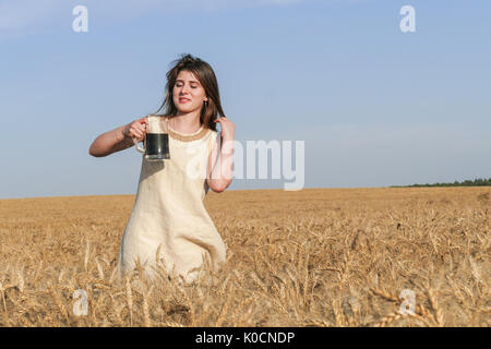 Junge attraktive Frau in wunderschönen natürlichen Kleid wandern mit mit einem Glas Bier in der goldene Weizen Feld während der Sonnenschein. Stockfoto