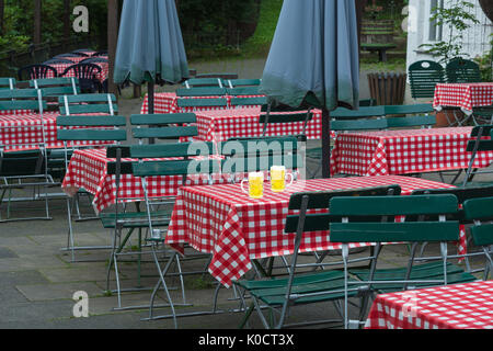 Zwei grosse Bierkrüge auf ein Bier Garten Tisch mit roten mit weißen Tischtüchern und grüne Stühle aus Holz in einem öffentlichen Biergarten. Stockfoto