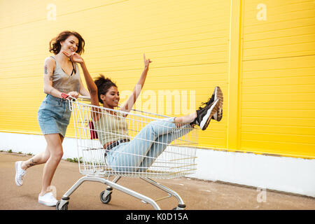 Bild der junge lächelnde Frauen Freunde über gelbe Wand. Viel Spaß mit Einkaufswagen. Stockfoto