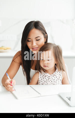 Bild von erstaunlichen jungen Mama am Tisch sitzen mit kleinen niedlichen asiatischen Mädchen zu Hause drinnen mit Laptop Computer schreiben Hinweise zum Notebook. Auf der Suche nach einem Stockfoto