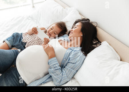 Gerne schwangere Frau und ihre kleine Tochter mit Handy beim zusammen Entspannen im Bett zu Hause Stockfoto