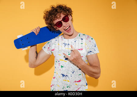 Stilvolle junger Mann in Sonnenbrille holding Skateboard und Kamera über Gelb Hintergrund isoliert Stockfoto
