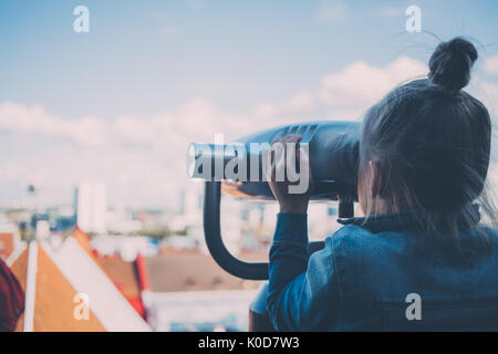 Junge Mädchen mit Blue Denim Jacken schaut münzbetriebene Fernglas auf der Aussichtsplattform, die Stadt und die Dächer auf dem Hintergrund. Stockfoto