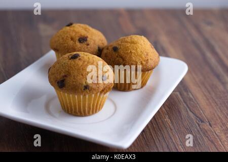 Drei miniatur Chocolate Chip Muffins auf weißem Rechteck, Platte und darkwood Tabelle Stockfoto