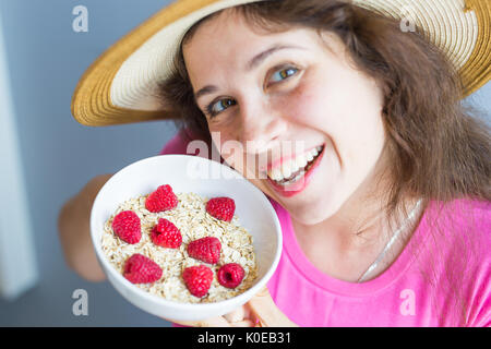 Lächelnde Frau halten gesund und natürlich Frühstück, Haferflocken und Himbeeren in eine Schüssel geben Stockfoto