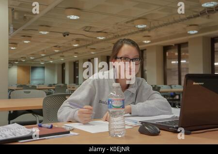 Eine Studentin, die Brille und Kopfhörer trägt, lächelt für ihr Foto, einen Bleistift in der Hand, während sie aus einem Notebook und Laptop, Papier und ihre Wasserflasche auf dem Tisch studiert, Sitz in einem leeren Raum auf EINER Ebene der Milton S Eisenhower Library an der Johns Hopkins University, Baltimore, Maryland, 2014. Mit Freundlicher Genehmigung Von Eric Chen. Stockfoto