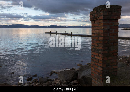 Ein See in der Abenddämmerung, mit einem alten Ziegel Spalte im Vordergrund und ein Pier an der Entfernung, mit warmen und sanften Farben in den Himmel und Wasser Stockfoto