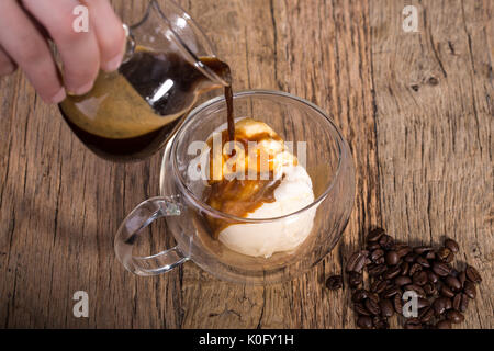 Gießen Kaffee Espresso in die Vanille Eis in eine doppelte Wand Glas auf einem Holztisch mit Kaffeebohnen Stockfoto