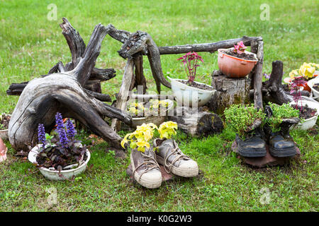 Florale Komposition mit Baumwurzeln, alte Schuhe, harte Hüte mit Blumen und Pflanzen. Ragged Objekte sind auf Erden Stockfoto