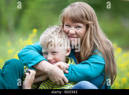 Junge Sohn ist in kräftigen Umarmungen, die glückliche Mutter, sitzen auf der Wiese im Park Stockfoto