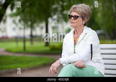 Alle Gedanken senior Europäerin konzentriert in Brillen. Sitzen auf der Bank im Park und schauen fern, Exemplar Stockfoto