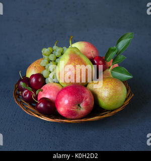 Herbst Früchte Mix - frische Äpfel, Birnen, Trauben und Pflaumen in einem wicker Holzplatte. Dunkelblauen Hintergrund. Ernte Konzept Stockfoto