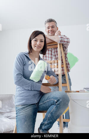 Glückliches Paar renovieren ihr Haus und Malerei ein Zimmer, sie lehnen sich auf einer Leiter und posieren mit Farbe rollen Stockfoto