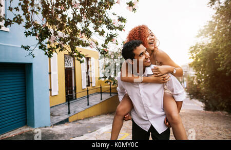 Lächelnden jungen Frau Huckepack reiten auf ihren Partner auf einer einsamen Straße. Junge Mann seinen Partner trägt auf seinem Rücken auf spielerische Stimmung. Stockfoto